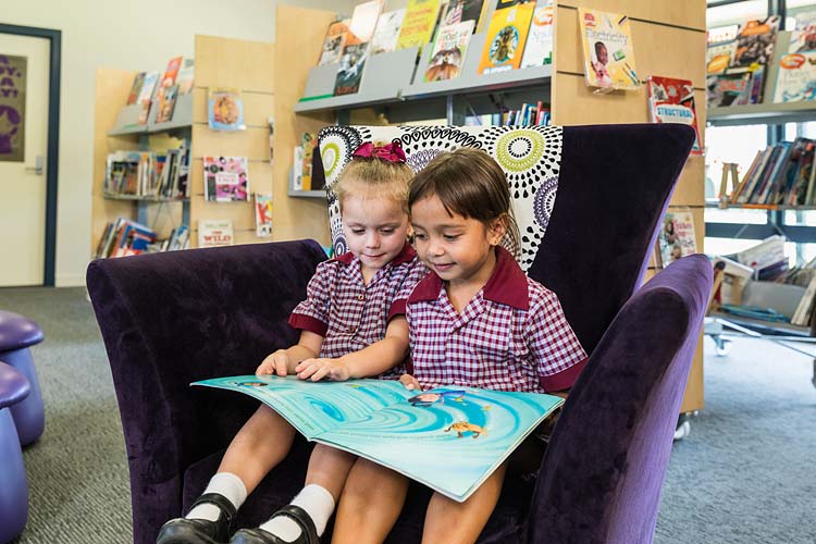 Two young school children sitting in library chair sharing a book