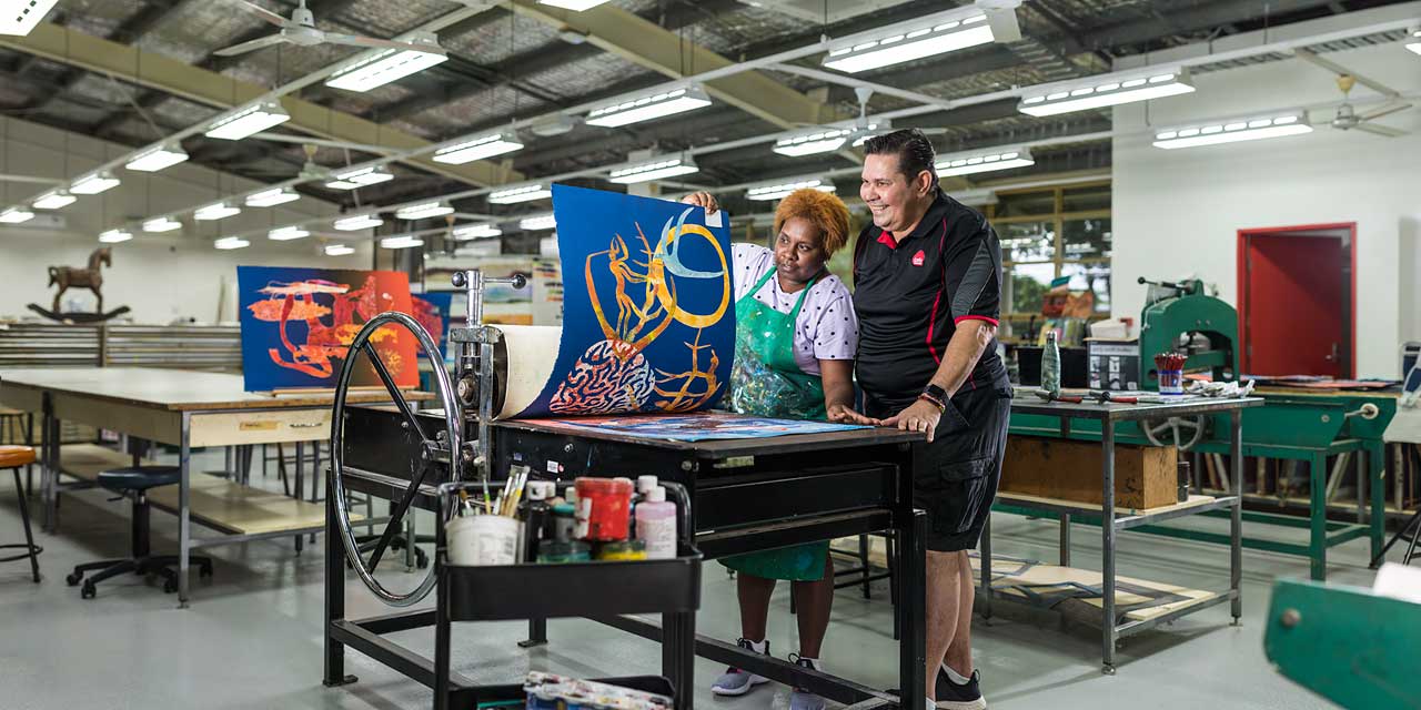 Indigenous arts student looking at artwork on printing press with teacher looking on
