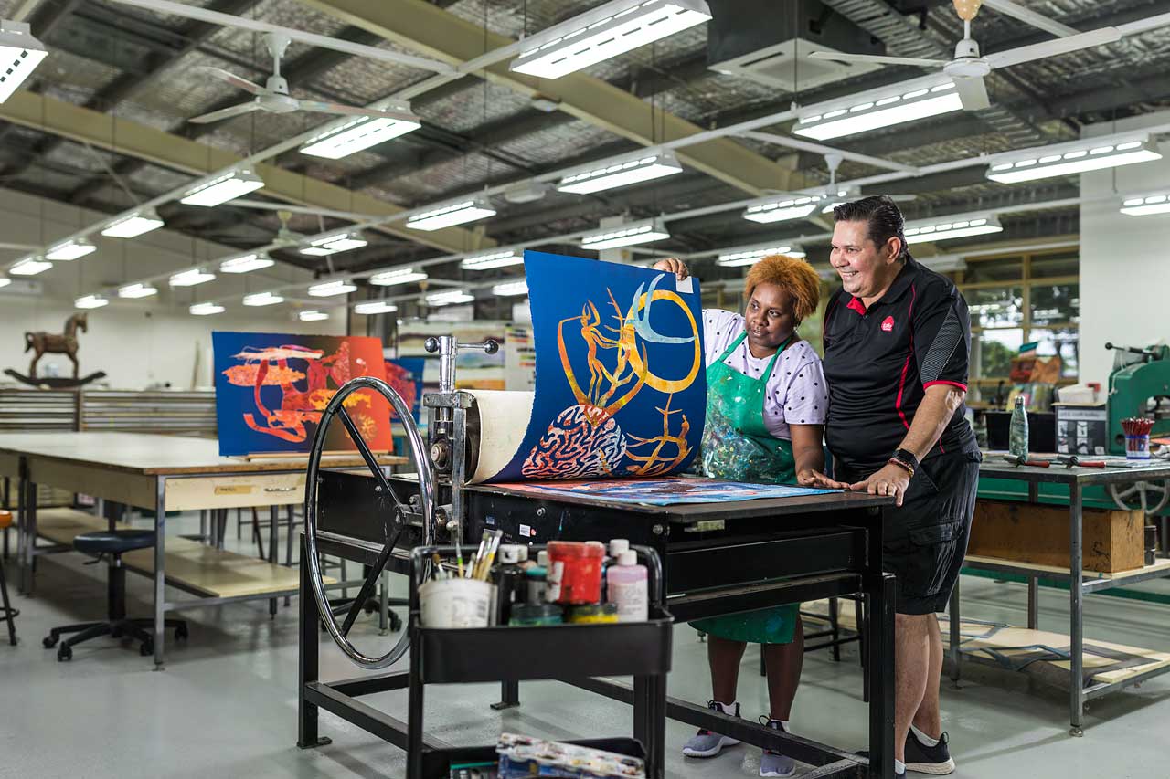 Torres Strait Islander student looking at artwork on printing press with teacher looking on