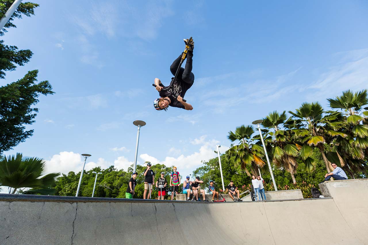 Ryan Williams of Nitro Circus upside down on a scooter - doing tricks at the Cairns skate park while young kids watch on