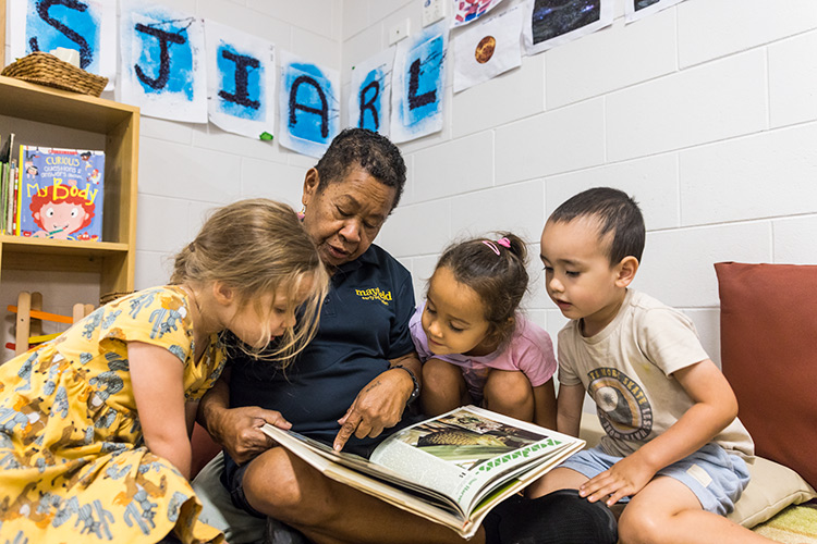 A female early learning educator surrounded by kids looking at a book