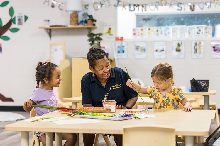 A female early learning educator watching on as kids enjoying painting using palm frond brushes