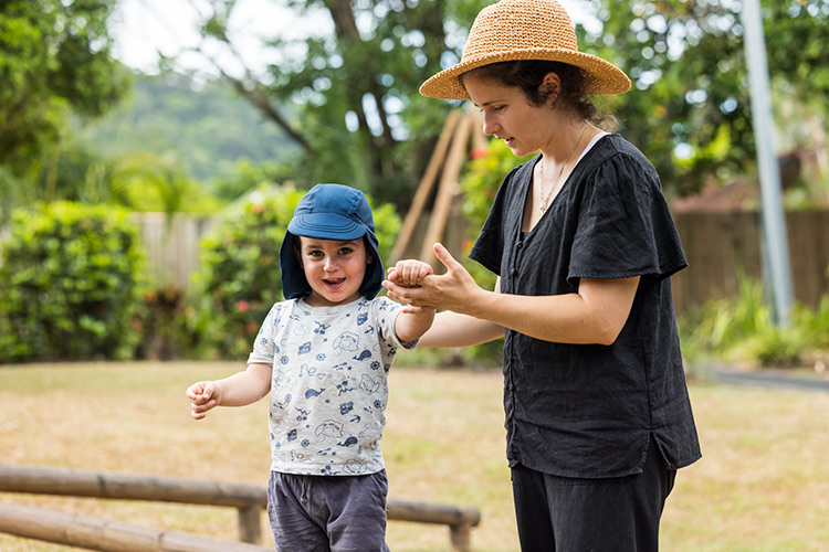 A child walking along a balance beam in childcare centre playground with help from an educator