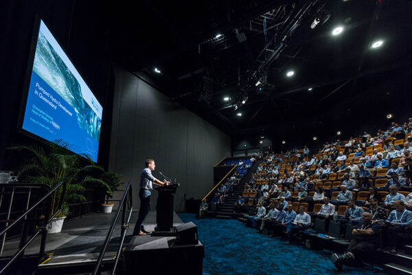 Male speaker at conference lecturn with audience looking on