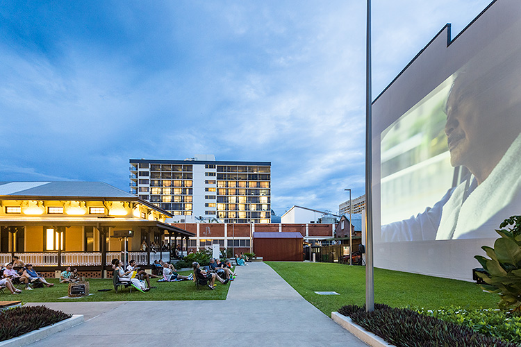 People seating on lawn of the Mulgrave Gallery watching a movie projected on a building wall